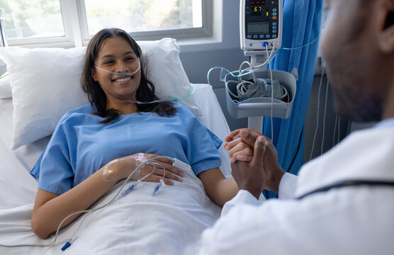 Happy diverse male doctor holding hand of female patient with iv drip in bed in hospital room