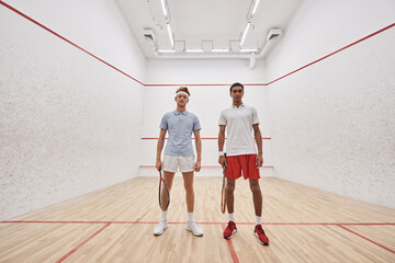 young interracial male friends in sportswear looking at camera and holding racquets for squash