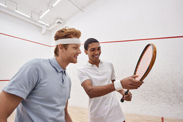 happy interracial sportsmen in active wear smiling and checking squash racquet inside of court