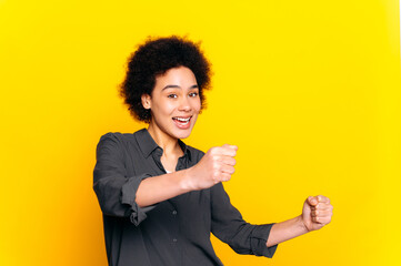 Cheerful attractive curly haired african american curly haired woman, holding in hands driving invisible car, imaginary steering wheel, stands on isolated yellow background, smiling, looks at camera