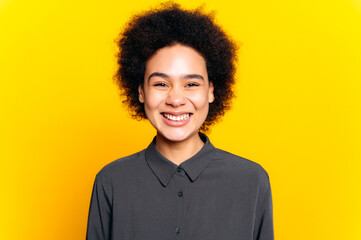 Obraz na płótnie Canvas Happy beautiful trendy african american or brazilian young woman with short curly hair, dressed in a shirt, looking at camera in a good mood, smiling friendly, standing on isolated yellow background