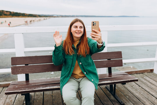 Happy 30s Women taking selfie and sitting on the bench outdoors. Young beautiful girl say Hi. Woman resting at the waterfront, seaside. High quality photo
