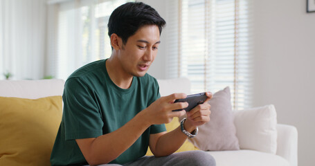 Asia people young man relax sitting easy at home sofa couch laugh smile enjoy play video game social media on mobile app, win online stream casino battle trade, leisure teen activity happy fun joy.