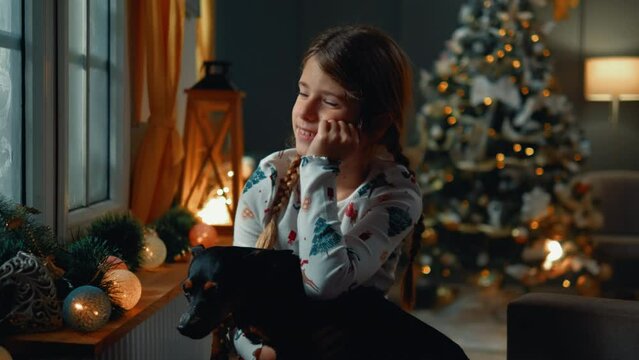 A little girl and her dog sit on the sofa and look out the window, they are waiting for Santa on Christmas or New Year's night
