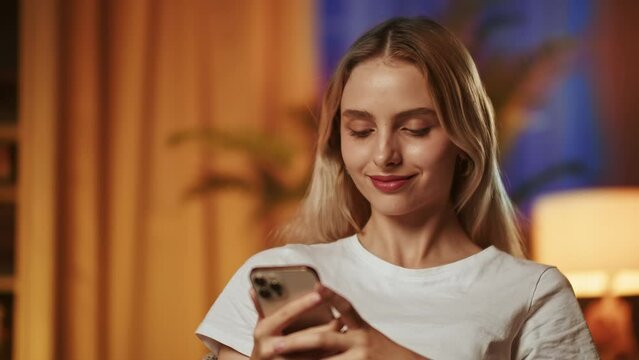 A smiling pretty blonde girl uses a smartphone, enjoys reading funny stories or jokes. A young lady with a happy satisfied face and a sly gloating gaze receives a notification about the punishment of
