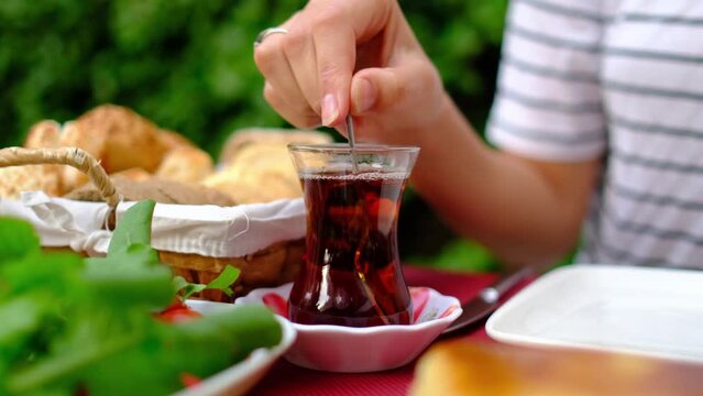 woman enjoys traditional Turkish breakfast on garden. She sits at table surrounded by greenery. woman stirring sugar in tea in a traditional Turkish tea cup called bardak