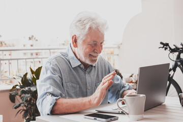 Video call concept. Portrait of smiling senior man sitting outdoor on a white table using laptop...