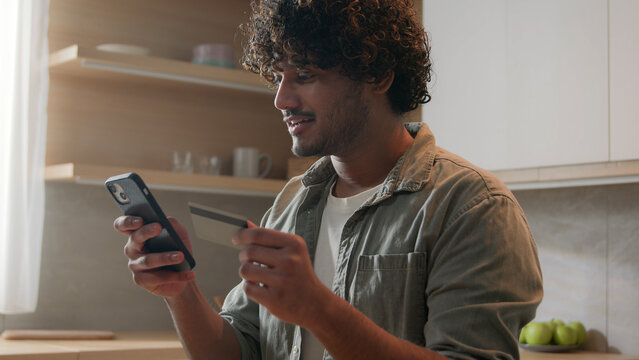 Happy Arabian Man With Phone And Credit Card Using Instant Mobile Payment At Home Kitchen Indian Guy Male Customer Shopper Buyer Buying Online Purchase Smartphone Shopping Food Delivery E-banking App