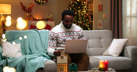 African American happy young man buying xmas gifts online on laptop paying with credit card on...