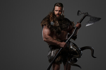 Fototapeta na wymiar A fierce bearded Viking warrior in fur and light armor, with a helmet attached to his belt, holding a large two-handed axe on a gray background