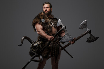 Fototapeta na wymiar A fierce bearded Viking warrior in fur and light armor, with a helmet attached to his belt, holding a large two-handed axe on a gray background