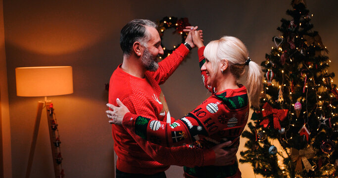Portrait of joyful Caucasian married middle-aged couple at decorated home celebrating New Year together at night. Wife and husband dancing near glowing xmas tree on Christmas. Love concept