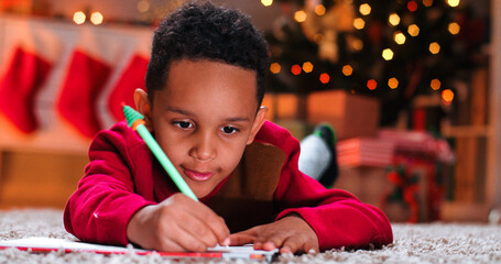 Close up of cute African American small boy drawing and writing on paper on floor in decorated home...