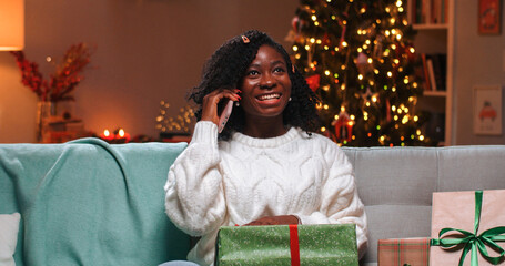 Close up portrait of happy African American pretty woman resting in decorated room on Christmas evening with xmas presents with glowing tree chatting on cellphone. Festive mood concept