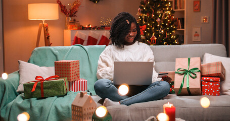 Joyful African American young pretty woman buying xmas presents online on laptop on xmas sales paying with credit card on New Year sitting on sofa in decorated room in evening with glowing tree