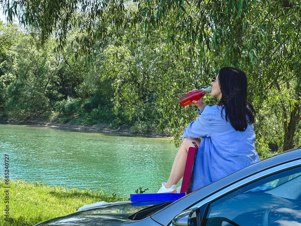 Poster happy woman sitting on car hood eating pizza looking on river