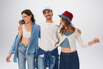 joyful young woman embracing woman in trendy casual attire and looking at camera on grey backdrop