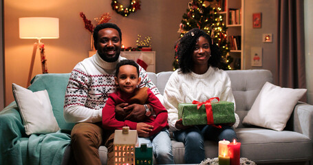 Portrait of happy African American family couple with cute small kid son sitting in decorated room at home with xmas gift, smiling to camera on Christmas. New Year's eve celebration concept