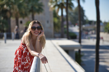 Obraz premium South American woman, young, beautiful, blonde with flower dress and sunglasses, leaning on a railing looking at camera smiling and happy. Concept travel, destinations, vacation.