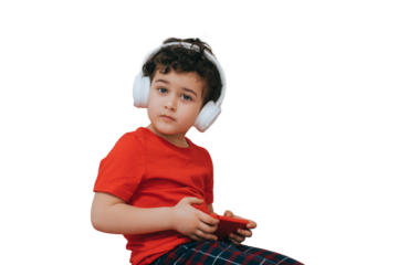 Cute little boy in headphones listening music looks at camera holds phone against transparent background with copy space. Handsome curly caucasian toddler playing games on phone.
