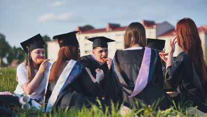 Graduates in black suits eating pizza in a city meadow.