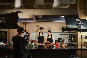 Ethnically diverse guy and girl working on culinary show production standing by kitchen counter in front of cameraman with technical equipment around - obrazy, fototapety, plakaty