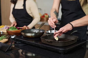 Closeup of unrecognizable man putting piece of butter on pan standing by kitchen stove with unknown...