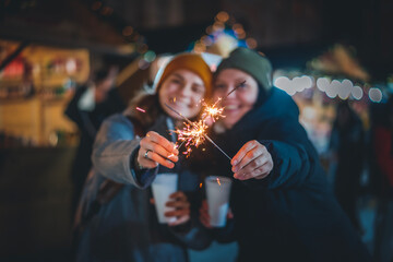 Two happy young women friends with mulled wine walking at the Christmas market with sparklers in...