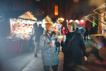 Two happy young women friends with mulled wine stands walking at a Christmas market in a European city