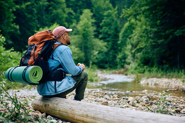 Rear view of hiker relaxing on tree trunk by mountain creek.