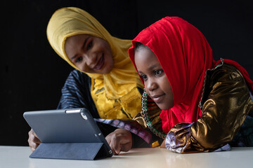 African Muslim girl doing her homework with digital tablet at home and her mother nearby, both wore colorful hijabs, online schooling concept on black background