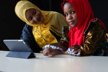 African Muslim girl doing her homework with digital tablet at home and her mother nearby, both wore colorful hijabs, online schooling concept on black background, girl looking at camera