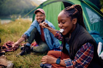 Happy black woman tasting roasted marshmallow while camping in nature.