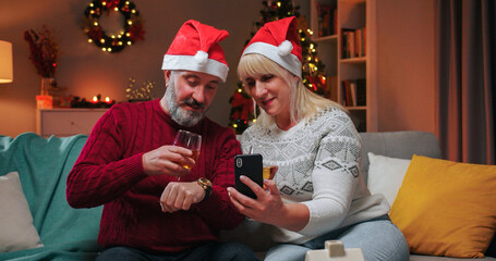 Portrait of joyful middle-aged Caucasian wife and husband in decorated room speaking on video on cellphone celebrating Christmas online with friends at home drinking wine New Year countdown