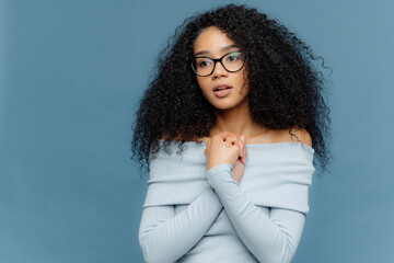 Serene young woman with glasses and curly hair, hands clasped, in thoughtful pose against a blue...