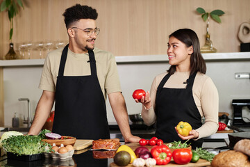 Young middle eastern guy and happy hispanic girl with vegetables in her hands looking at each other while posing at kitchen - Powered by Adobe