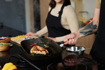 Closeup of unrecognizable man holding pan with prepared piece of meat showing it to camera with unknown woman standing on background, focus on meat