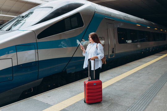 Young redhead woman waiting train with backpack and using smart phone. Railroad transport concept, Traveler. 30s Woman with suitcase walking at railroad station platform. Travel to vacation by train