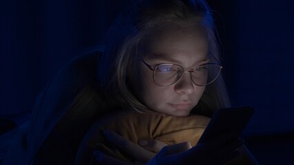 Young woman with glasses is texting on her smartphone lying in bed at home at night close up....