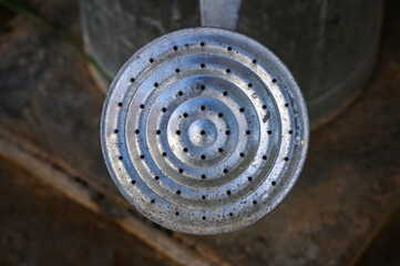 "Close-up of the round head of an aluminum metal watering can. In the foreground, the holes from which the water emerges, with a blurred background of muddy ground where the garden watering can is pla
