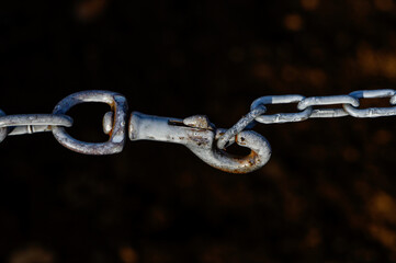 "A close-up of the metal hook of a dog leash attached to an old chain with aged and whitened links against a dark background."