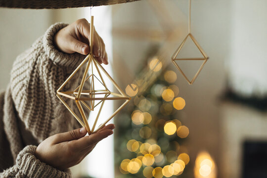 Hands In Cozy Sweater Hanging Rustic Nordic Straw Decoration On Background Of Festive Golden Lights Bokeh In Modern Farmhouse Living Room. Decorating Home With Christmas Straw Spiders.