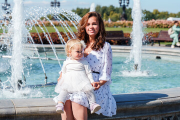 Mom and daughter of baby toddler laugh and hug near the fountain in the park on a summer day