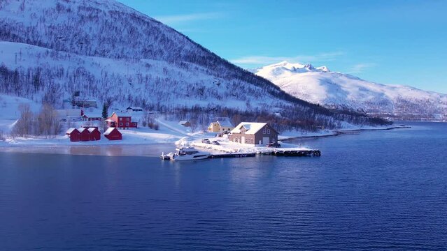 Snow covered fishing village on coastline in winter, Norway. Surroundings of town Tromso. Panoramic aerial view landscape of nordic mountains, houses, rorbu, boats and ocean. Troms county, Fjordgard