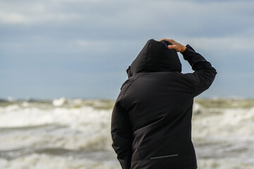 A man in a black jacket with a hood holds the hood and looks at the stormy sea, rear view © Zigmunds