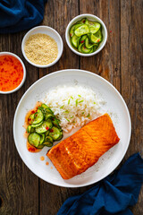 Seared salmon steak with boiled white rice and sliced cucumber on wooden table
