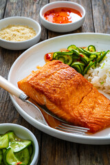 Seared salmon steak with boiled white rice and sliced cucumber on wooden table
