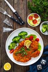 Crispy breaded fried chicken cutlet with boiled broccoli and fresh vegetables on wooden table
