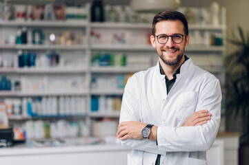 Picture of a young happy pharmacist standing with arms crossed and smiling at the camera.