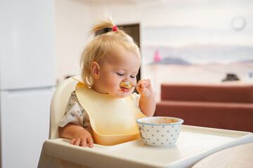 Little toddler girl eats herself with a spoon in a bib in the kitchen